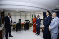 Foto de cinco pessoas em pé, sendo quatro homens e uma mulher. Ao lado esquerdo da foto, um homem lendo o documento que está na prancheta em que segura. Ao lado direito da foto, a mulher e os outros quatro homens olhando para frente, prestando atenção ao que se é lido.