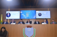 Mesa de honra composta por 7 homens sentados. Brasão da Câmara Municipal de São Luís de forma centralizada à mesa. Ao fundo, três telas de televisores com imagens voltadas ao homenageado.