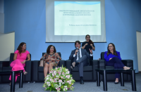 Foto de um homem e de quatro mulheres num auditório. O homem e três mulheres estão sentados e a outra mulher está em pé. O auditório tem parede azul, onde tem um projetor. À frente, no piso do auditório, há um arranjo floral.