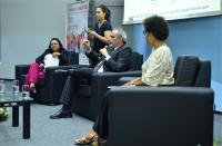 Foto tirada em auditório. Três pessoas sentadas em poltronas de cor preta, sendo duas mulheres aos extremos e um homem sentado ao centro. Ao fundo, uma intérprete de libras em pé.