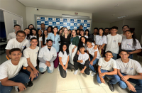 Grupo de pessoas posando para foto. Alguns estão posicionados em pé e outros abaixados. Ao centro, em destaque, três mulheres, a primeira vestida com uma blusa na cor azul e calça na cor vinho, a segunda vestida com roupa preta e a terceira com blusa verde e calça branca; ladeadas de instrutor e de alunos e alunas vestidos com uniforme de cor branca