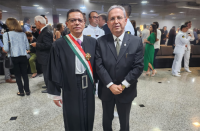 Dois homens posicionados em pé, posando para foto, ambos usando óculos de grau.  O homem da esquerda usa toga na cor preta e faixa transpassada listrada nas cores verde, branca e vermelha, com a comenda. Ao fundo, algumas pessoas em pé circulando pelo ambiente.