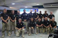 Foto de uma mulher e vários homens; alguns estão em pé e outros, agachados. Todos vestem calças na cor cáqui e camisas pretas. Alguns dos homens estão usando óculos.