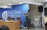 Foto de uma mulher em pé que está gesticulando com as mãos. Ela usa roupas nas cores verde claro e pretas e está com o cabelo preso num coque. A mulher está ao lado de uma mesa e cadeiras e próxima a uma parede azul onde está escrito Escola Judicial na cor branca.