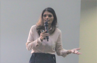 Foto de mulher clara, com cabelos escuros, usando blusa de mangas compridas em tom de rosa claro e saia preta. Ela fala ao microfone, que segura com a mão direita, e gesticula com a mão esquerda. Ao fundo, vê-se parede em tons claros.