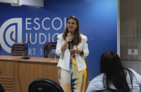 Foto de uma mulher que está em pé, falando ao microfone. Ela veste macacão colorido e um blazer branco. Na frente dela, há uma mulher sentada. Atrás dela, há uma mesa com cadeiras, e uma parede azul onde tem uma logomarca e está escrito Escola Judicial na cor branca.