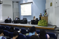  Quatro pessoas fazendo a abertura de um seminário dentro de um auditório e uma intérprete de libras. 