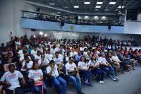 Foto de vários homens e mulheres, entre os quais, jovens aprendizes, sentados e em pé, em um amplo auditório