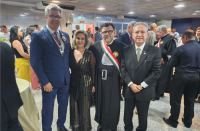 Grupo de quatro pessoas posando para a foto, posicionados em pé, sendo três homens e uma mulher. Dois homens vestidos com terno, um homem vestido com toga e uma mulher usando vestido preto com detalhes dourados. Ao fundo, algumas pessoas em pé circulando pelo ambiente. 