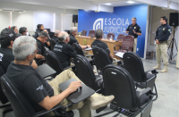 Uma mulher e alguns homens sentados em cadeiras na cor preta; dois homens em pé, um deles próximo a uma mesa com cadeiras e em frente a uma parede azul onde está escrito Escola Judicial; o outro está mais à direita. Todos vestem calças cáqui e camisas pretas.