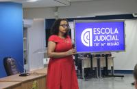 Foto de uma mulher que está em pé; ela tem cabelos encaracolados, usa óculos, está com vestido vermelho e segura um microfone com as duas mãos. À direita, projetor de imagem onde está escrito Escola Judicial, na cor branca.