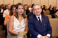 Duas pessoas sentadas lado a lado, em poltronas claras, em um evento que acontece dentro de um auditório, posando para a foto. A imagem referida se trata de uma mulher (ao lado esquerdo) e de um homem.