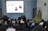 Foto de uma mulher falando numa tribuna diante de um auditório composto por mulheres e homens. Na parede central do auditório, um projetor de imagens. Na parte superior da parede branca, à direita, o brasão do Poder Judiciário. Atrás da tribuna, três bandeiras oficiais.
