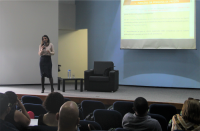 Foto panorâmica da instrutora do curso no palco, próxima a uma poltrona preta e pequena mesa lateral. No canto superior direito, vê-se projeção de slides. Na parte de baixo, plateia de costas.