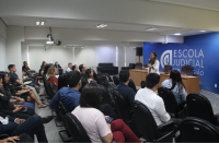 Foto de uma mulher que está em pé num auditório, falando ao microfone para uma plateia composta por mulheres e homens que estão sentados