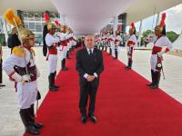 Foto de um homem claro, usando terno escuro, de pé sobre tapete vermelho na entrada do STF e ladeado por guardas usando o fardamento oficial dos Dragões da Independência.