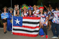 Grupo de oito mulheres posando para foto e segurando a bandeira do Estado do Maranhão (nove listras horizontais intercaladas em três cores: vermelho, branco e preto), e no lado superior esquerdo da bandeira, um quadrado azul com uma estrela na cor branca). 