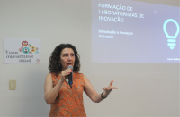 Foto de uma mulher branca, de cabelos encaracolados, usando uma blusa laranja, falando ao microfone, que segura com a mão direita, e gesticula com a mão esquerda. Ela está à frente de uma parede branca, na qual se vê um cartaz à sua direita e uma projeção de slides à sua esquerda.