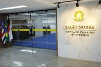 Foto da entrada do Salão Nobre, com bandeiras do Maranhão, Brasil e do TRT-16 hasteadas ao lado esquerdo, uma porta vidro e, na parede, na parte superior, ao lado direito, um brasão da república dourado e o nome SALÃO NOBRE JUSTIÇA DO TRABALHO TRT 16ª REGIÃO com letras em dourado. 