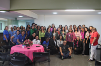 Foto de mulheres e homens em uma sala ampla. Parte das pessoas está em pé, outras estão agachadas e um homem está sentado. No ambiente há uma mesa coberta por uma toalha rosa. Ainda sobre a mesa um arranjo floral na cor rosa, além de celulares.