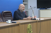 Foto de um homem branco que está sentado numa cadeira marrom, ele está em frente a uma bancada e está falando ao microfone. Na bancada de madeira, há um brasão do Poder Judiciário.