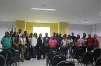 Foto de várias mulheres e vários homens que estão em pé num auditório. Estão em frente a uma parede amarela e branca.