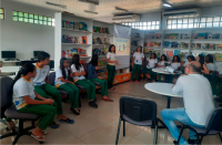 Foto de vários estudantes que estão numa biblioteca e um de homem que está de costas. Duas jovens estão em pé e as outras pessoas estão sentadas. Na sala, há vários livros, revistas e um banner pendurado em uma das estantes.