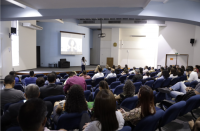 Foto de várias pessoas (homens e mulheres) sentadas num auditório participando de seminário; no destaque, palestrante. Na parede central do auditório, um projetor com as informações do evento. À direita, o brasão do Judiciário na parte superior da parede.