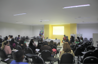 Foto de mulheres e homens num auditório. Todos estão sentados e, em frente a uma parede amarela, há uma mesa coberta por uma toalha branca, e duas mulheres à direita que estão debatendo, observadas por um homem, que está no lado esquerdo, e por pessoas que estão na plateia. No ambiente, há também bandeiras oficiais e banners.