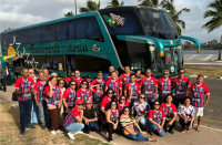 Foto de vários homens e mulheres sentados e em pé; a maioria usa camisa com listras verticais e uma parte lisa, nas cores vermelha, azul, branca e preta. Todos estão em frente a um ônibus na cor verde, com detalhes nas cores preta, branca e amarela.