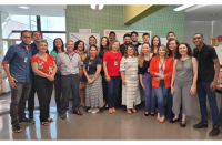 Foto de vários homens e mulheres de pé e sorrindo, à frente de uma parede verde.