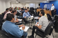 Foto de uma turma em sala de aula, dividida em grupos de estudo, sendo observada pelo professor, de pé à frente dos alunos.