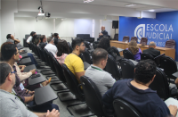 Foto de homens e mulheres sentados em cadeiras pretas. Todos estão num auditório assistindo a um homem que está falando ao microfone. Numa parede central pintada de azul, está escrito Escola Judicial, na cor branca, ao lado da logomarca da Escola. 