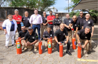 Foto de homens e mulheres num espaço aberto, alguns em pé e outros agachados. Também há extintores de incêndio que estão próximos aos homens que estão agachados.