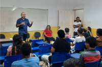 Foto de várias pessoas sentadas, entre elas, estudantes, e de dois homens que estão em pé. Todos estão numa sala de aula. Um dos homens está falando e está em frente a um quadro branco que está pendurado na parede.