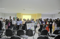 Foto de várias mulheres e de homens num auditório. Todos estão em pé. Parte está em frente a uma parede amarela. Duas das mulheres estão segurando um banner.