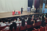 Foto de várias pessoas (homens e mulheres) num auditório, a maioria está sentada em cadeiras vermelhas, na plateia, e cinco pessoas estão em pé no palco. Um dos homens que está em pé veste blazer cinza e calça azul marinho e está falando ao microfone. No palco também há uma mesa coberta com toalha vermelha e cadeiras e um banner.