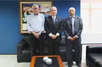 Três homens posando para foto, dentro de uma sala com parede de fundo na cor azul, mesa de centro em madeira com objetos decorativos em formato esférico..