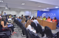 Foto de homens e mulheres sentados em um auditório, que estão assistindo a uma mulher que está em pé e falando ao microfone, em frente a uma parede azul, onde está escrito Escola Judicial. A mulher tem cabelos compridos e escuros e está vestindo um macacão verde. 