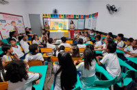 Foto de vários estudantes e três mulheres adultas que estão numa sala de aula. Das três mulheres, duas estão em pé, em frente aos estudantes e a outra está sentadas. Os estudantes do ensino fundamental estão sentados em cadeiras amarelas e verdes. Nas paredes, quadros e adesivos com as letras do alfabeto.