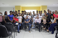 Foto de vários estudantes (moças e rapazes), além de mulheres e homens adultos num auditório. Alguns em pé e outros sentados formando um semicírculo.