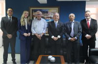 Seis pessoas posando para foto, dentro de uma sala com parede de fundo na cor azul. Cinco homens e uma mulher. A frente, mesa de centro em madeira com objetos decorativos em formato esférico.