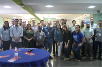 Foto de homens e mulheres que estão em pé num salão, em frente a uma mesa redonda que está coberta por uma toalha azul. Sobre a mesa, panfletos, placa de papel  e um microfone.