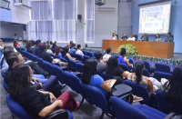 Foto de várias pessoas (homens e mulheres) num auditório. Parte das pessoas está sentada em cadeiras azuis e a outra parte está sentada em cadeiras colocadas junto a uma ampla mesa, no palco do auditório. Atrás do palco, uma tela de projeção. As paredes do auditório são pintadas nas cores azul e cinza. Também tem no ambiente aparelhos de ar condicionado e cortinas de persianas.