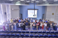 Foto de várias pessoas (homens e mulheres) que estão em pé num auditório. Algumas pessoas estão sobre o palco. Outras, na frente do palco. Algumas mulheres e alguns homens estão segurando uma cartilha. Atrás, na parede central pintada de azul, há uma tela de projeção. As paredes do auditório são pintadas nas cores azul e cinza. Também tem no ambiente aparelhos de ar condicionado e cortinas de persianas. Na frente das pessoas, várias cadeiras azuis.