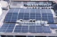 Foto de Foto de várias placas solares fotovoltaicas que estão localizadas no topo de um edifício. Também se vê na imagem telhados cobertos com telha brasilit, além de condensadores de ar condicionado.