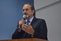 Foto de homem claro e grisalho, falando ao microfone, que segura com a mão direita,  e gesticulando com a mão esquerda. Ele usa terno escuro e camisa clara, Ao fundo parede azul e branca.