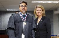 Duas pessoas posando para foto dentro de um auditório. Um homem à esquerda, vestido com toga, e uma mulher vestida com blaser preto.