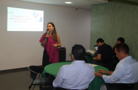 Foto de uma mulher clara, de cabelos longos e escuros, em pé, falando ao microfone, que segura com a mão direita. Ela usa blusa rosa e calça preta e está  à frente de uma parede branca, onde se projeta uma apresentação de slides. Na plateia, veem-se pessoas de costas, em mesas circulares, assistindo à palestra.