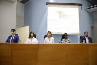 Mesa de honra composta por cinco pessoas, três mulheres ao centro e dois homens nas extremidades. A foto foi tirada dentro de um auditório com paredes azul e branca.